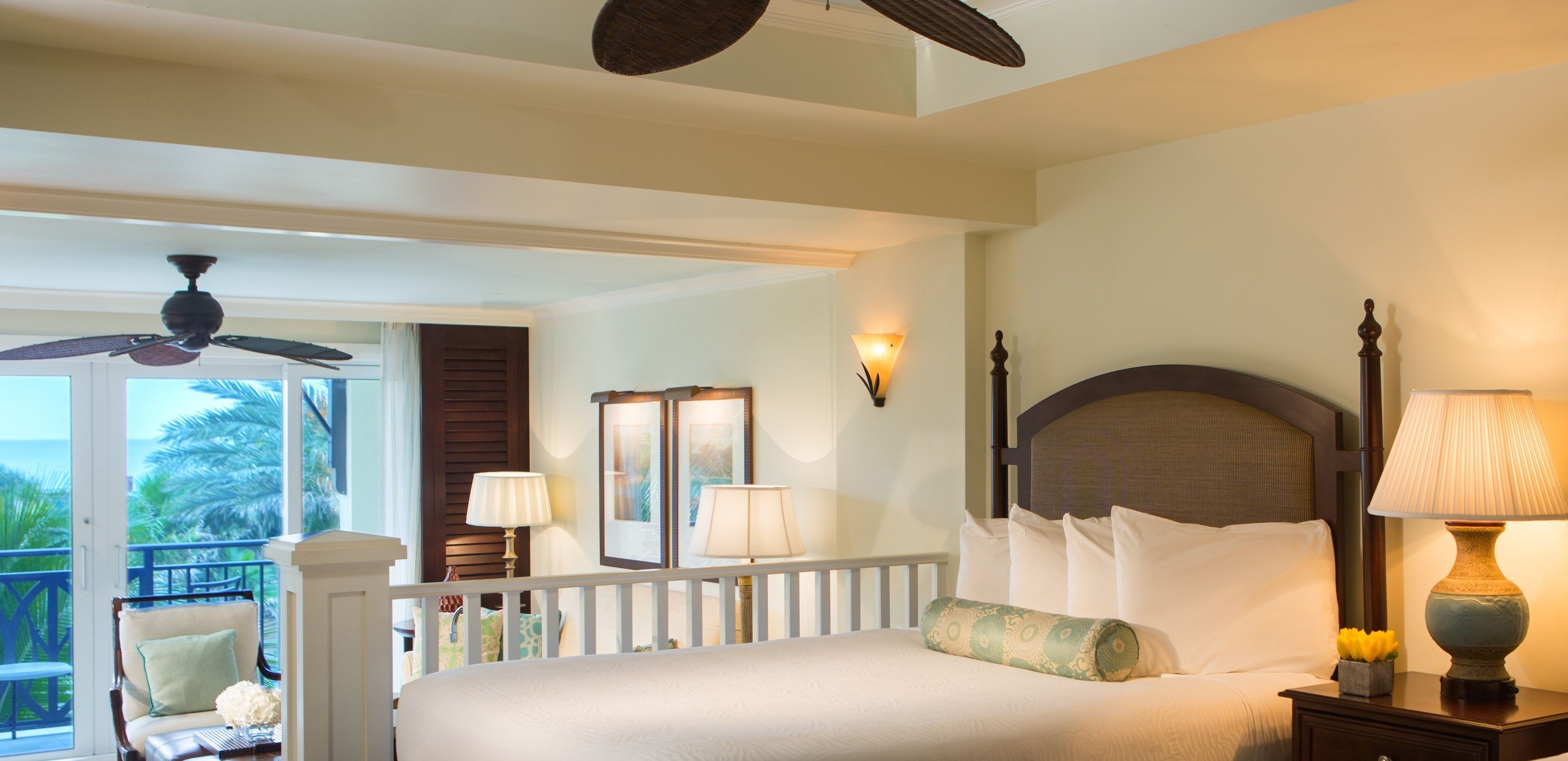 A hotel room with a bed, two bedside lamps, a ceiling fan, a balcony with a chair, and a view of palm trees.