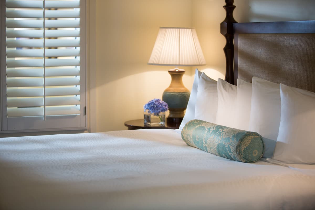 A neatly made bed with white linens, a decorative pillow, a bedside lamp, and a vase with flowers on a nightstand.