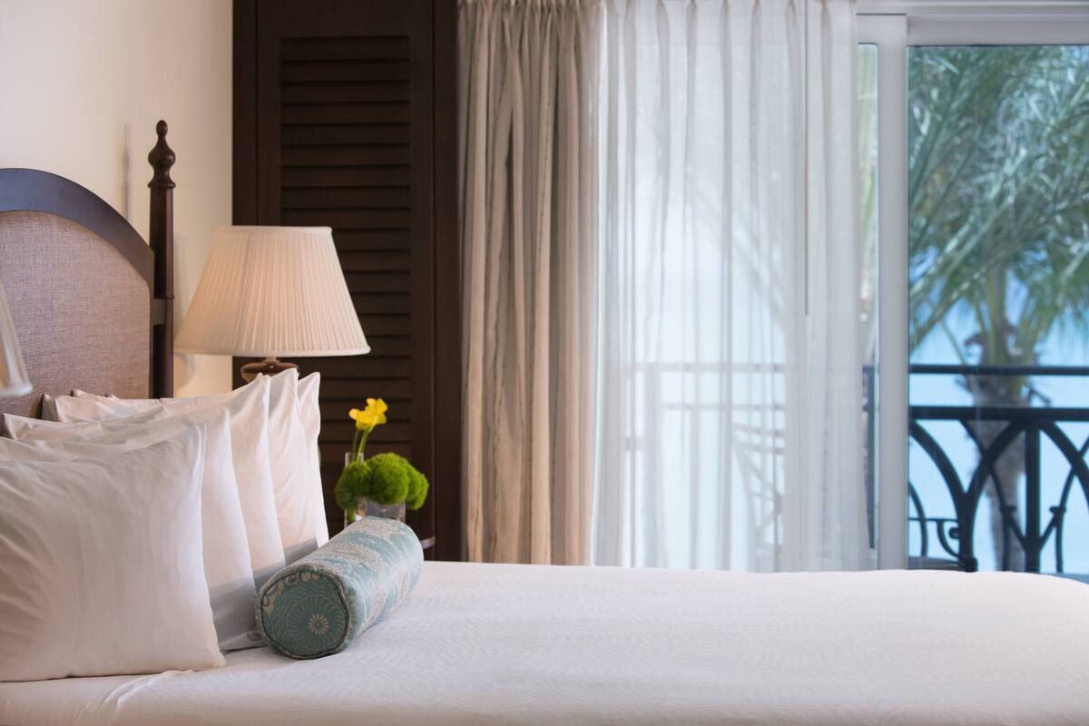 A neatly made bed with white linens and a decorative pillow. A lamp is positioned beside the bed, and a small vase with a yellow flower is on the nightstand.