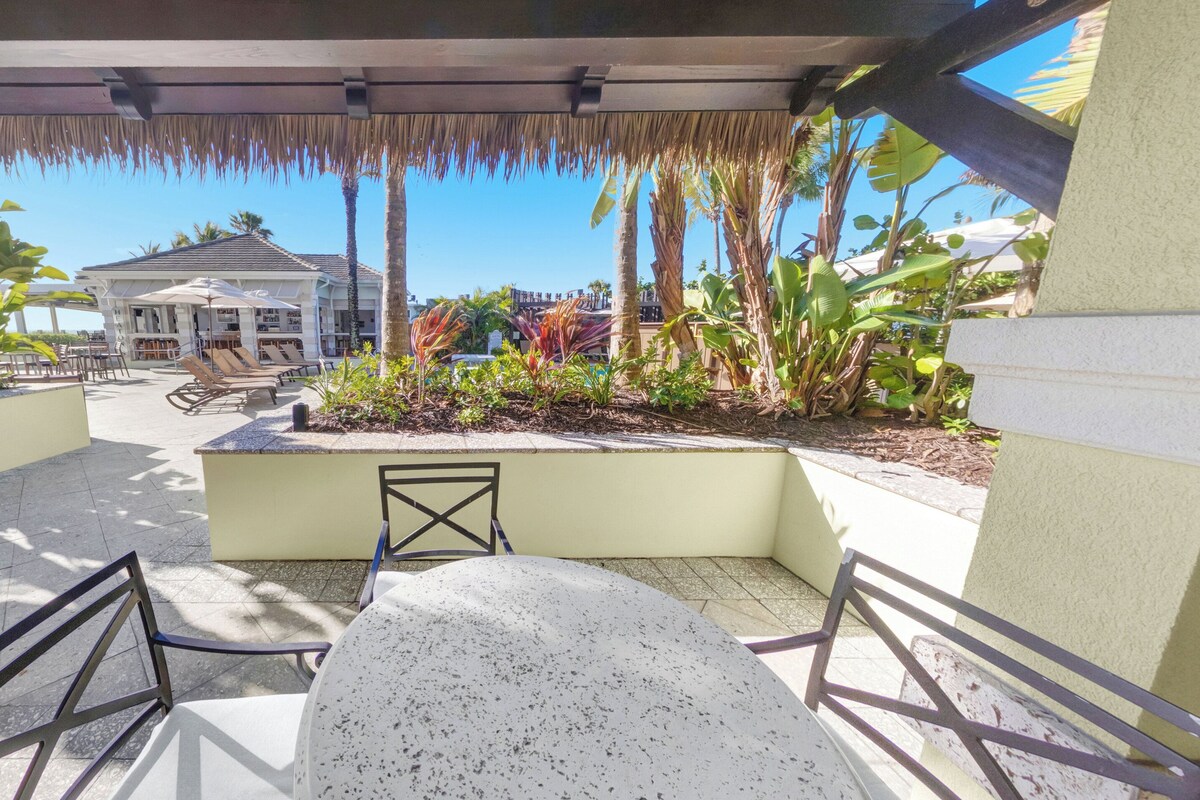 Un espace extérieur aménagé, abrité et doté d’une table et de chaises, entouré de plantes tropicales, avec un bâtiment visible en arrière-plan.