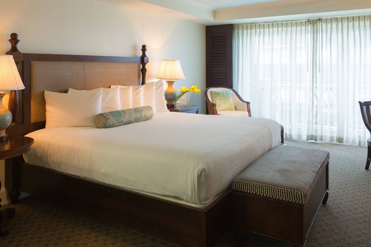 A large bed with multiple pillows, a nightstand with a lamp, a chair, and a window with curtains.