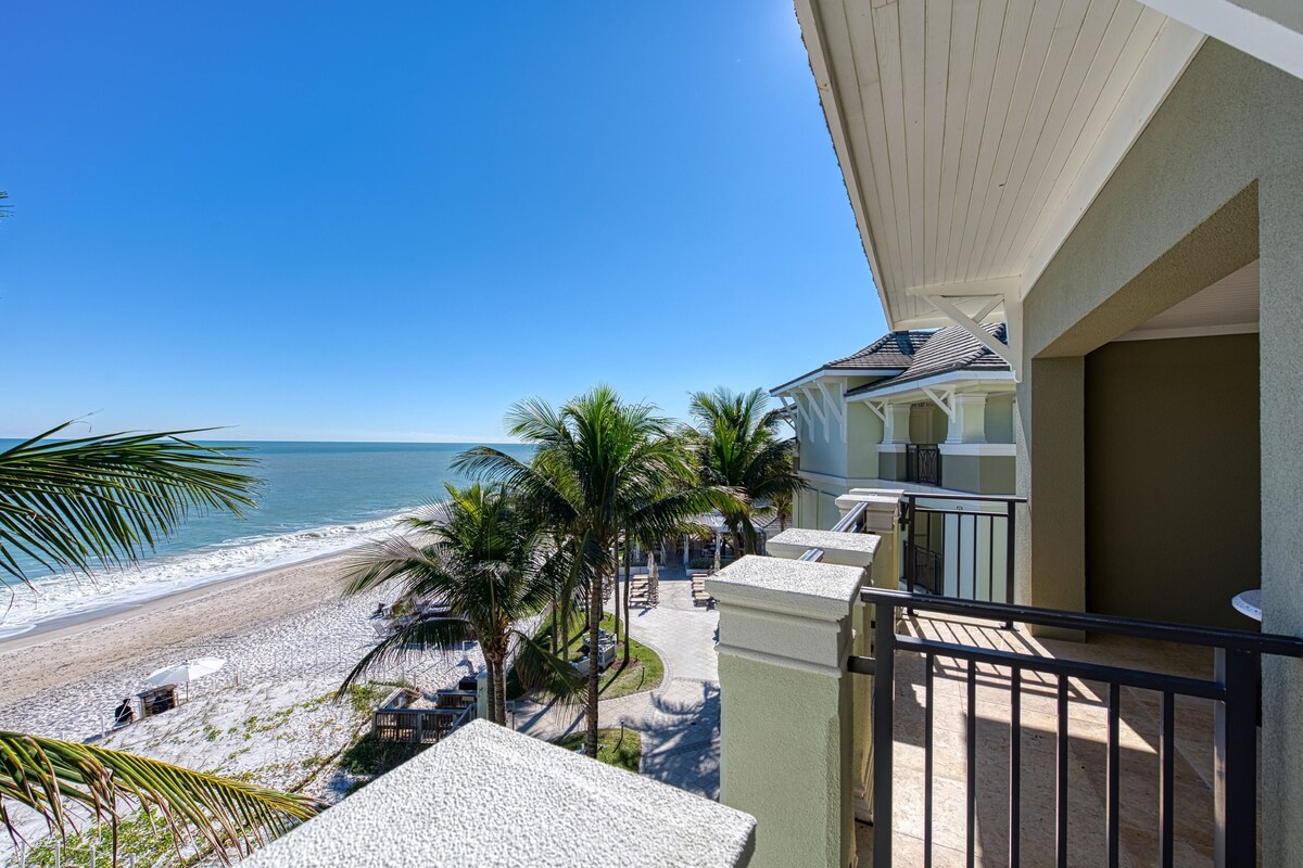 Une propriété située en bord de mer, dotée d’un balcon offrant une vue sur la plage, les palmiers et l’océan.