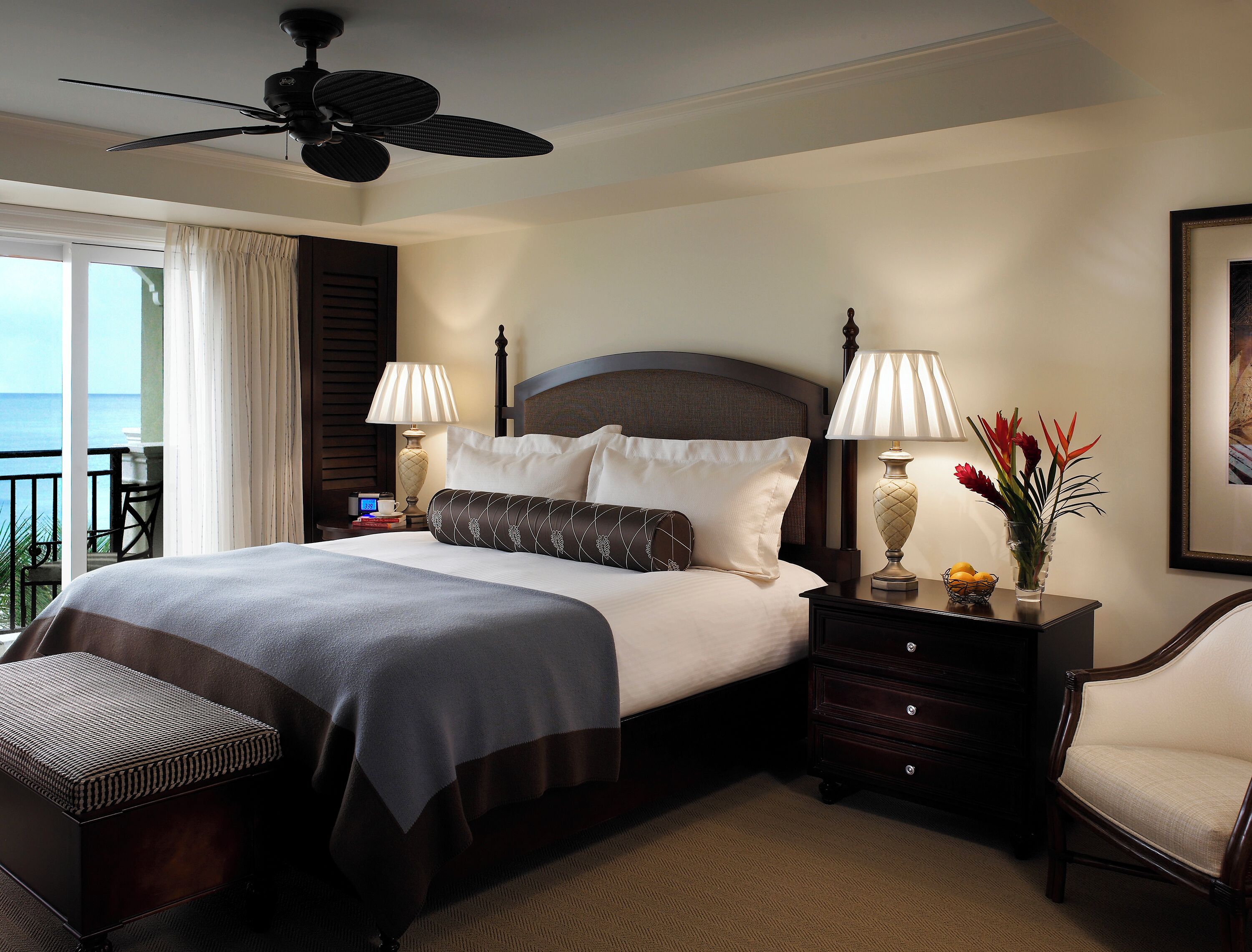 A hotel room with a bed, two bedside lamps, a nightstand, a chair, a small table with a vase of flowers, a ceiling fan, and a view of the sea through a window.