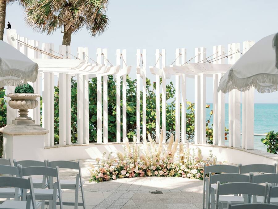 A white pergola with a white fence, chairs arranged for seating, and a view of the ocean.