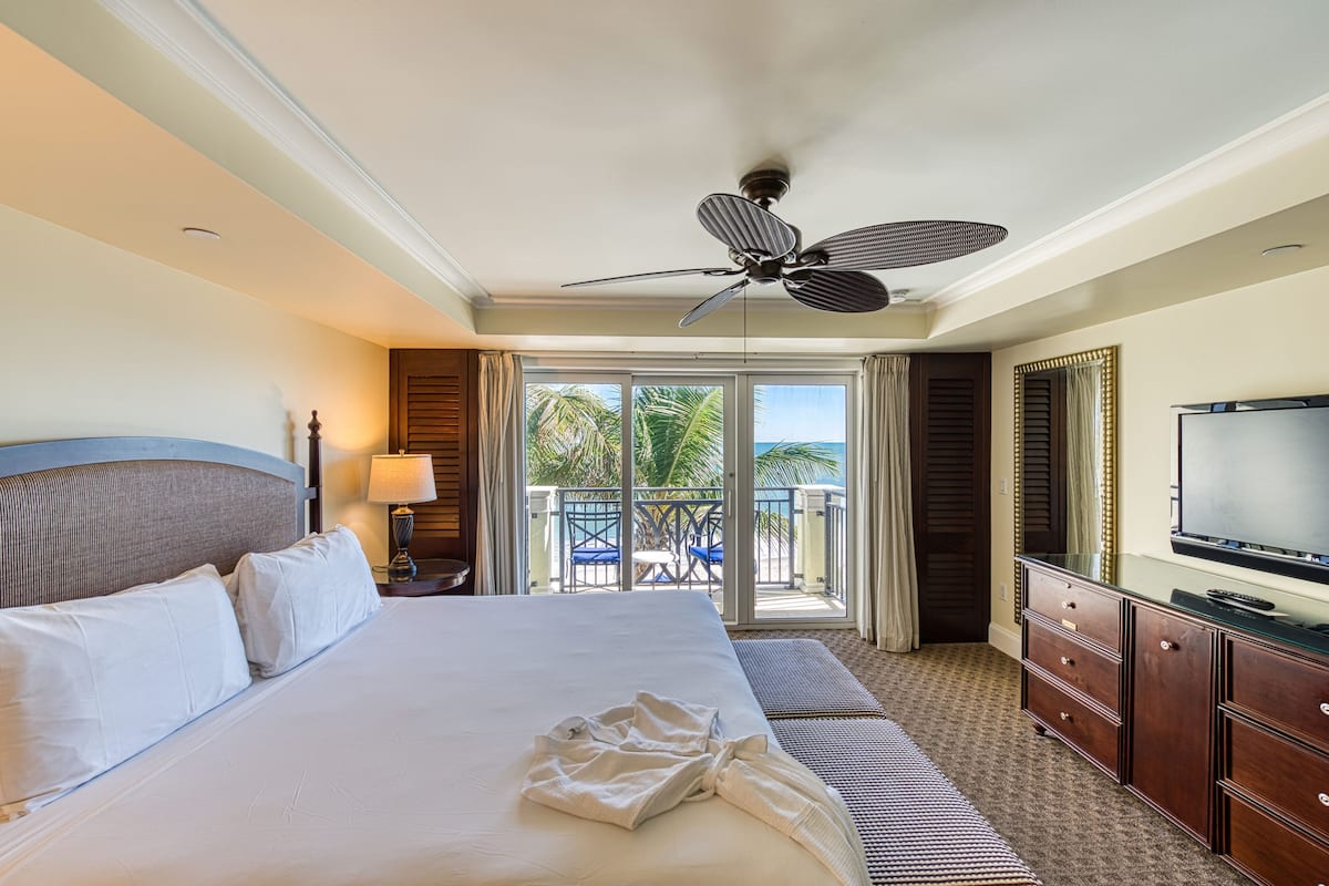 A hotel room with a large bed, a ceiling fan, a TV, a balcony with a view of palm trees, and a sliding door.