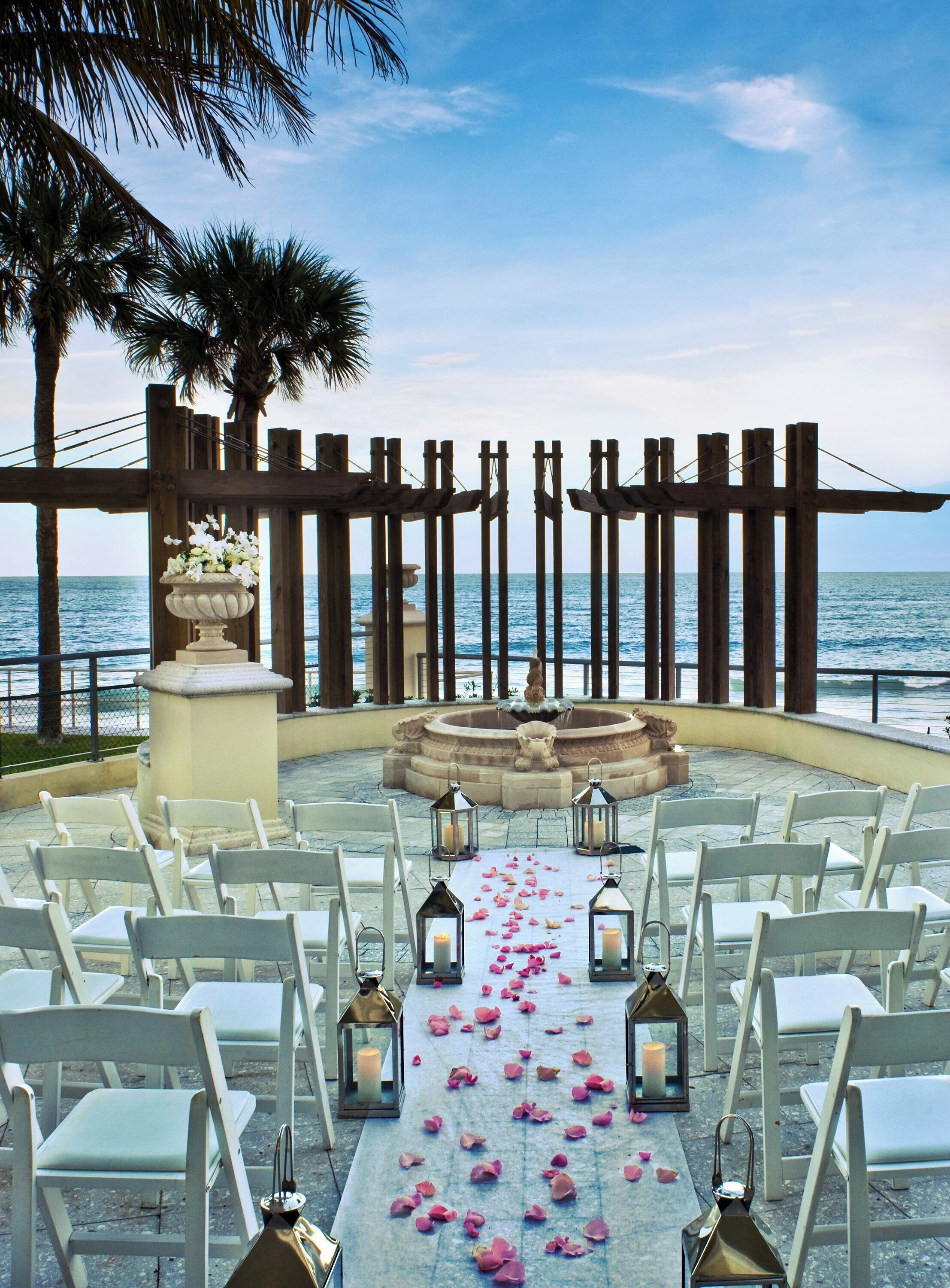 An outdoor wedding ceremony setup with white chairs arranged in rows, a floral aisle, and a fountain centerpiece.