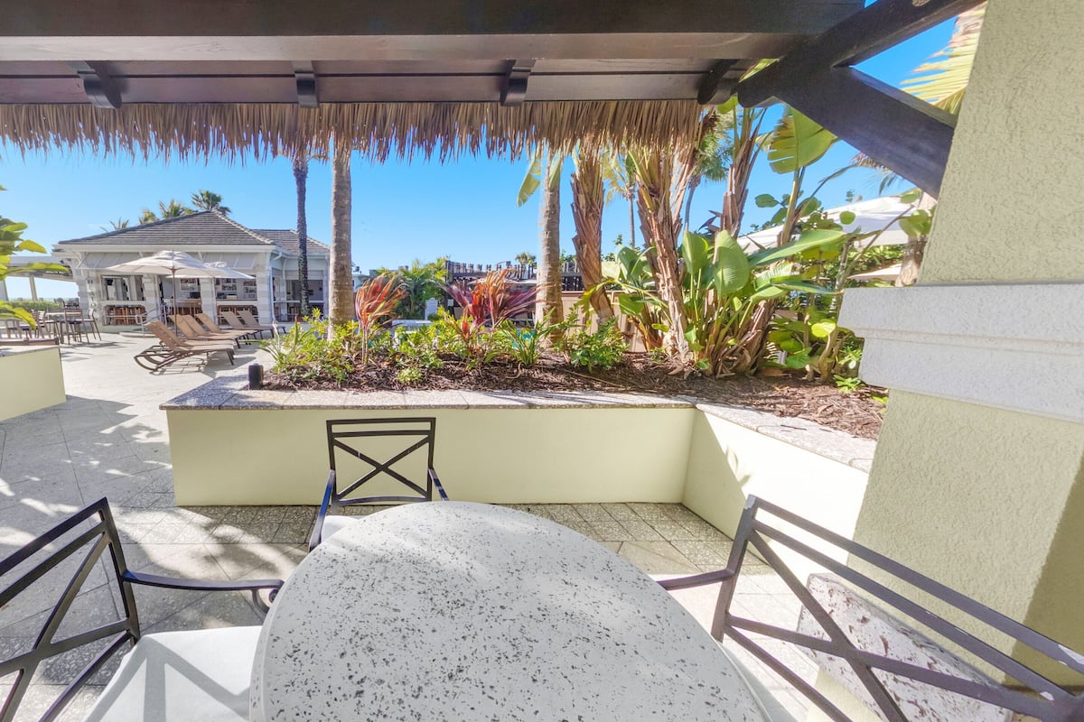 A covered outdoor seating area with a table and chairs, surrounded by tropical plants and a building in the background.