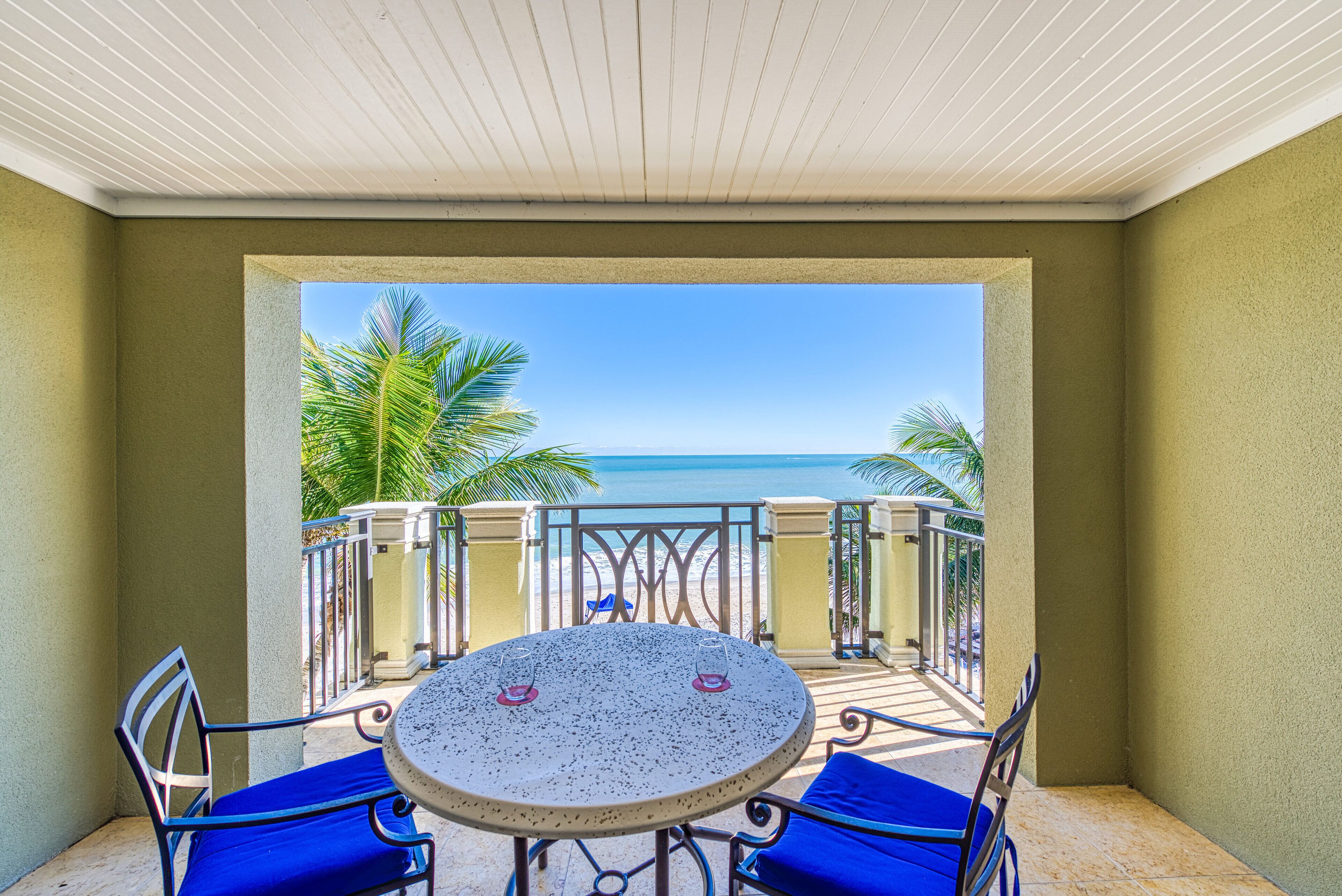 Un balcon avec une table ronde et des chaises bleues, donnant sur une plage bordée de palmiers et sur l’océan.