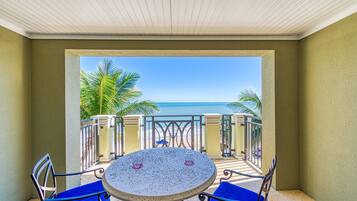 Un balcon avec une table ronde et des chaises bleues, donnant sur une plage bordée de palmiers et sur l’océan.