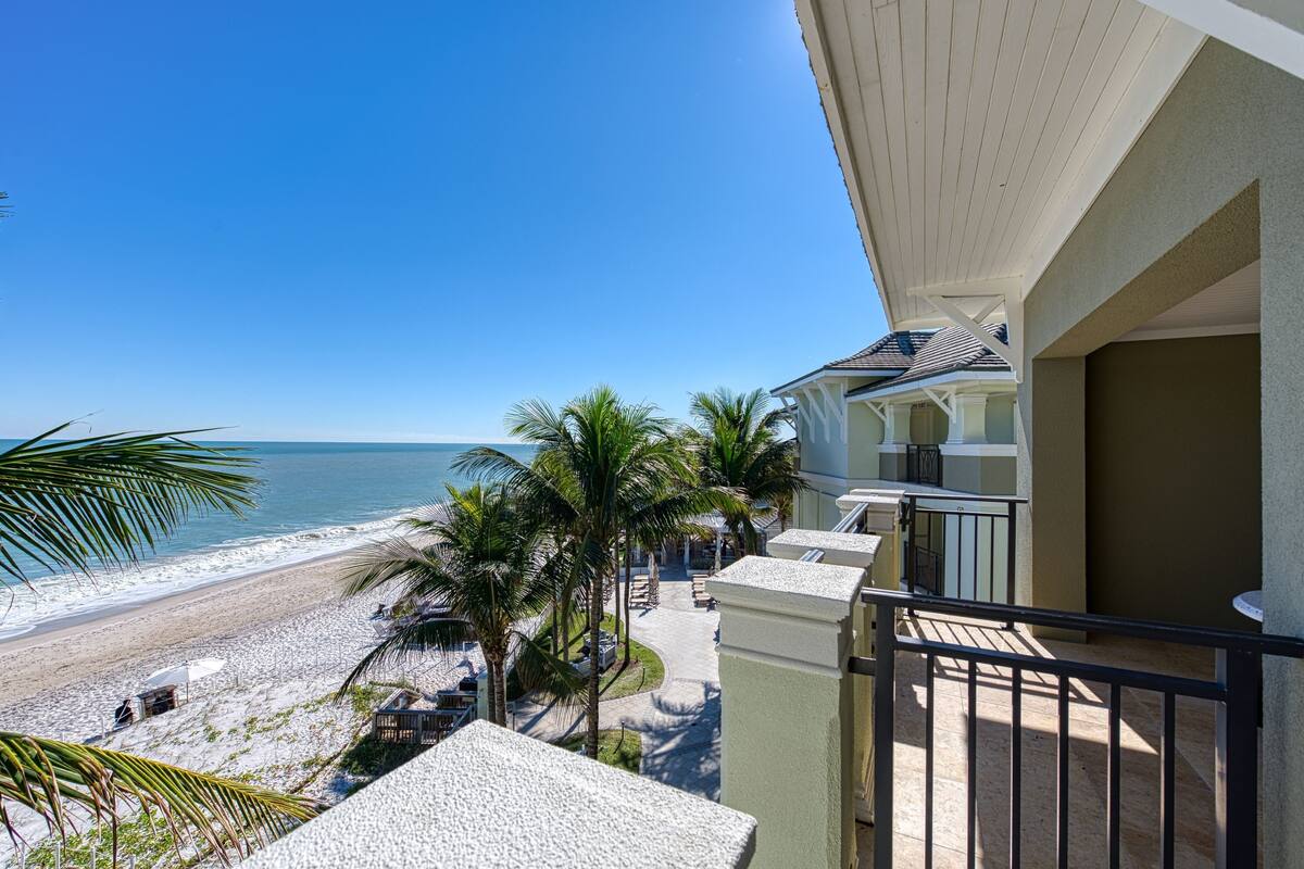 A beachfront property with a balcony overlooking the beach, palm trees, and a clear view of the ocean.