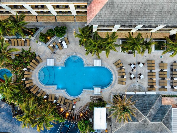 Vue aérienne d’un complexe hôtelier doté d’une piscine centrale, de plusieurs espaces de détente avec des chaises longues et entouré de palmiers.