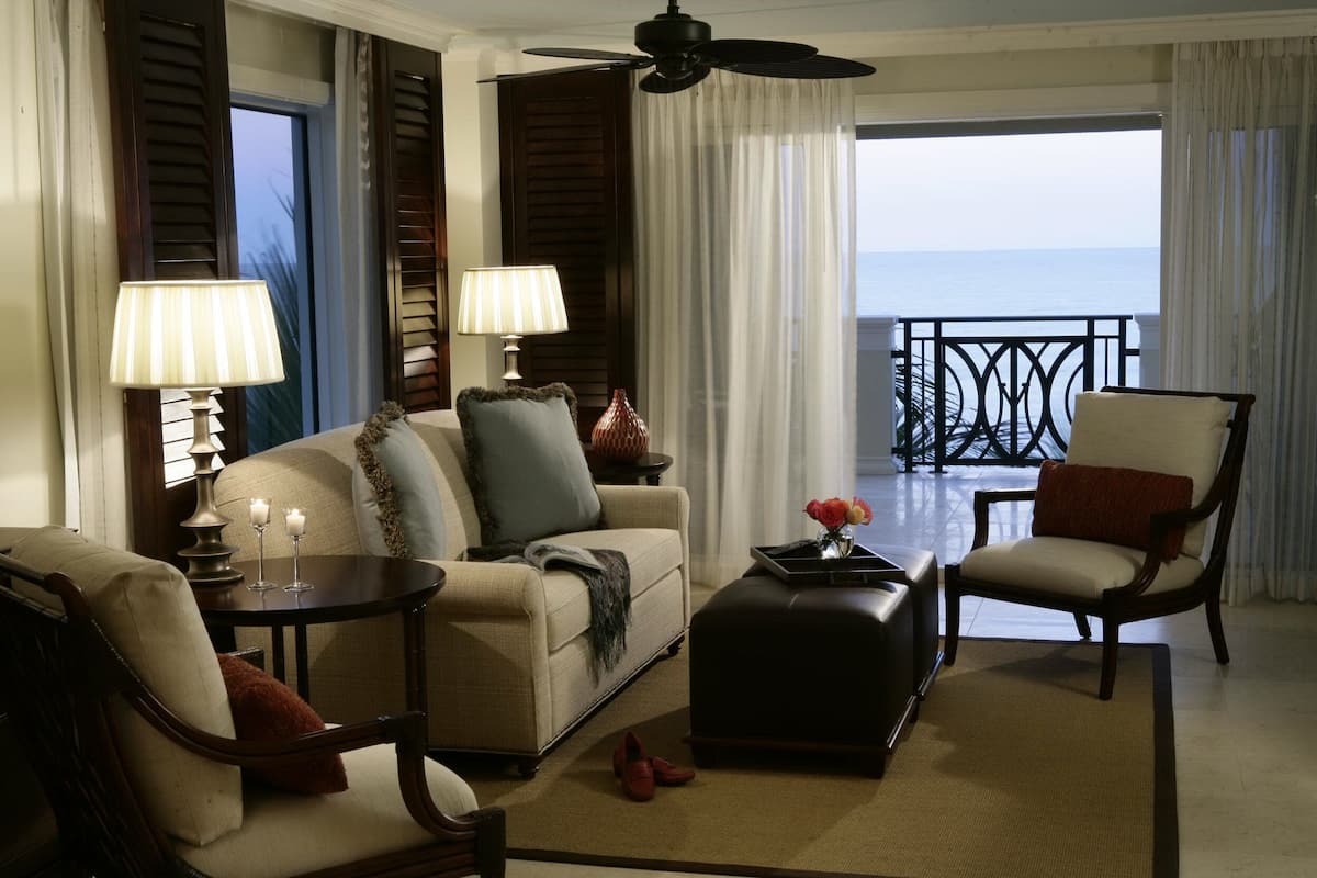 A well-lit living room with a sofa, armchairs, a coffee table, and lamps. There is a balcony with a railing and a view of the ocean.