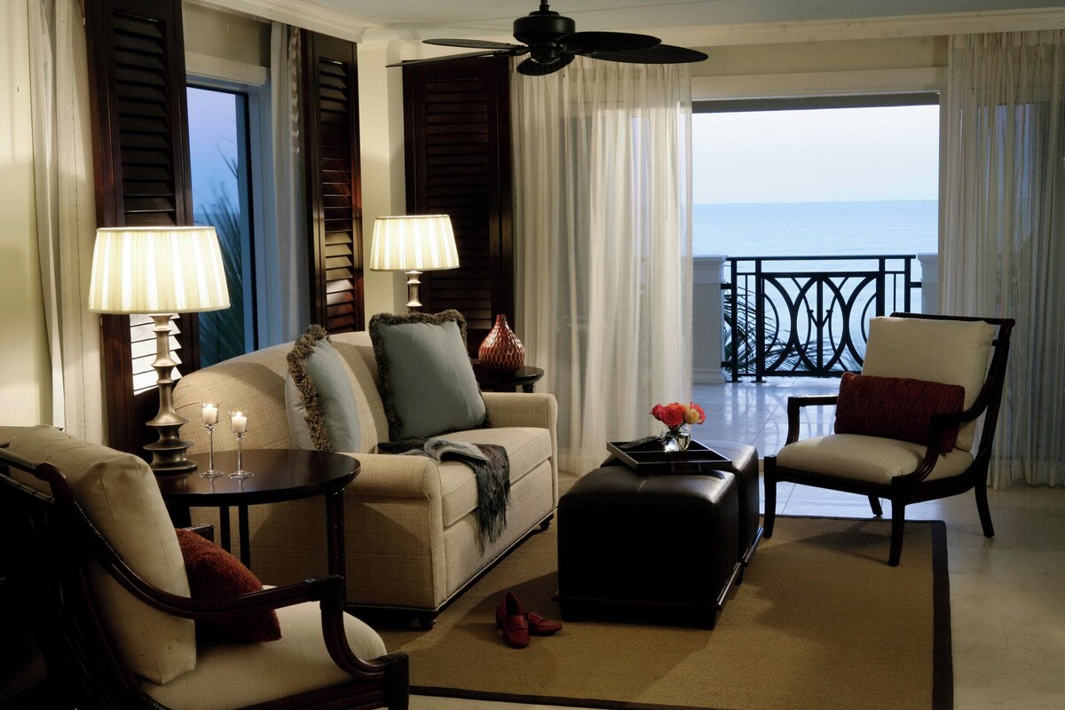 Un salon bien éclairé, avec un canapé, des fauteuils, une table basse et des lampes. Il y a un balcon avec une balustrade et une vue sur l’océan.