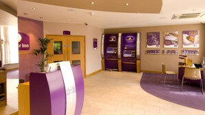 A hotel reception area with a reception desk, a plant, a wooden door, and a purple carpet. - Premier Inn London Kensington - Olympia (London)