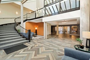 A spacious lobby with a modern staircase, a herringbone floor pattern, and a seating area with a lamp and a plant.