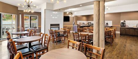 A spacious dining area with multiple round tables and wooden chairs, a kitchen in the background, and a large wooden column.