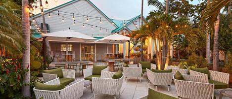 An outdoor seating area with wicker furniture and umbrellas, surrounded by palm trees and a building with a white exterior.