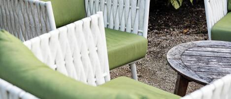 A white wicker chair with green cushions, a round wooden table, and a background of dense green foliage.