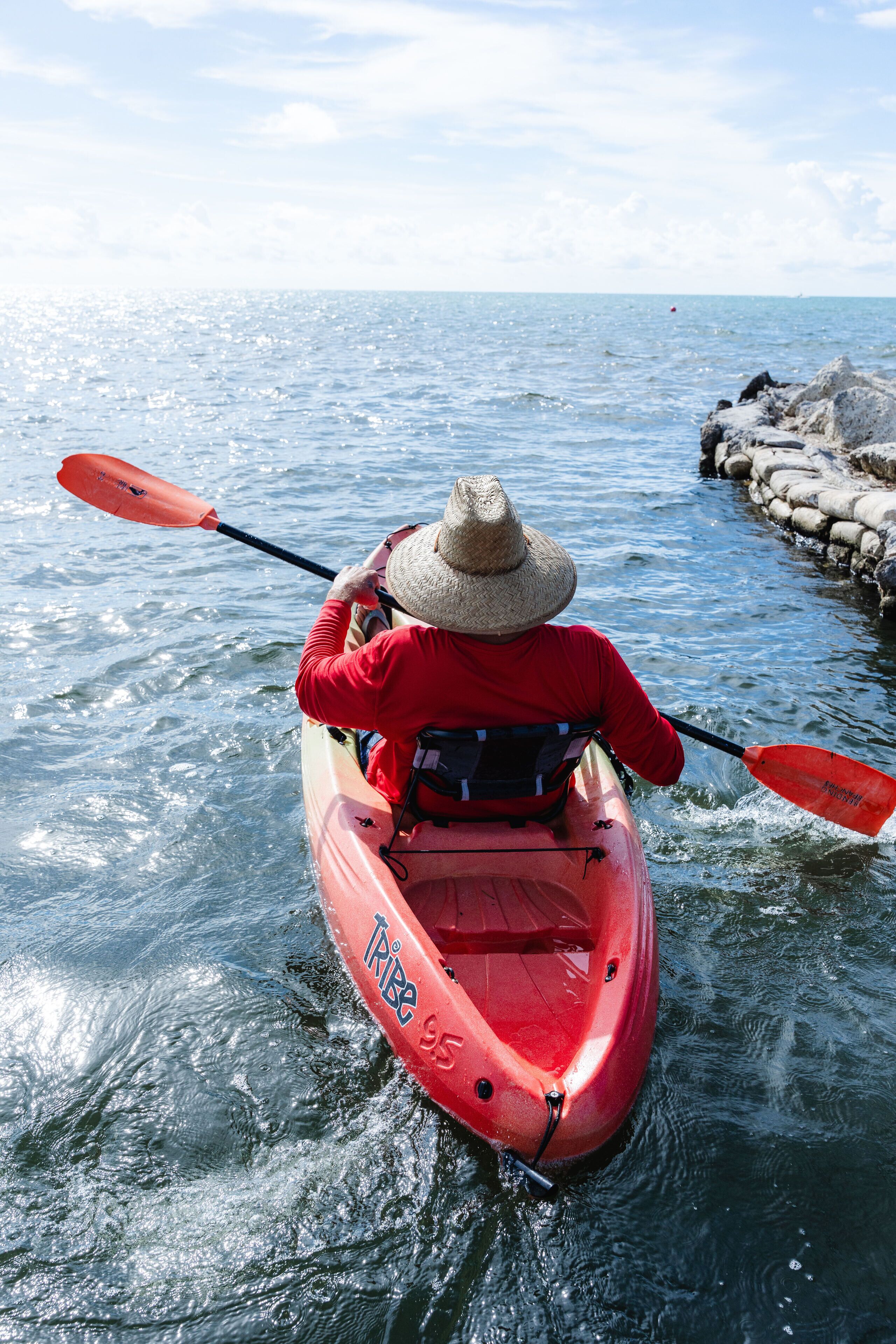 Una persona practica kayak en un río, usando un kayak rojo y un sombrero de paja.