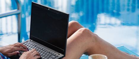 Una persona usando una computadora portátil junto a una piscina, con un vaso de jugo de naranja sobre una mesita.