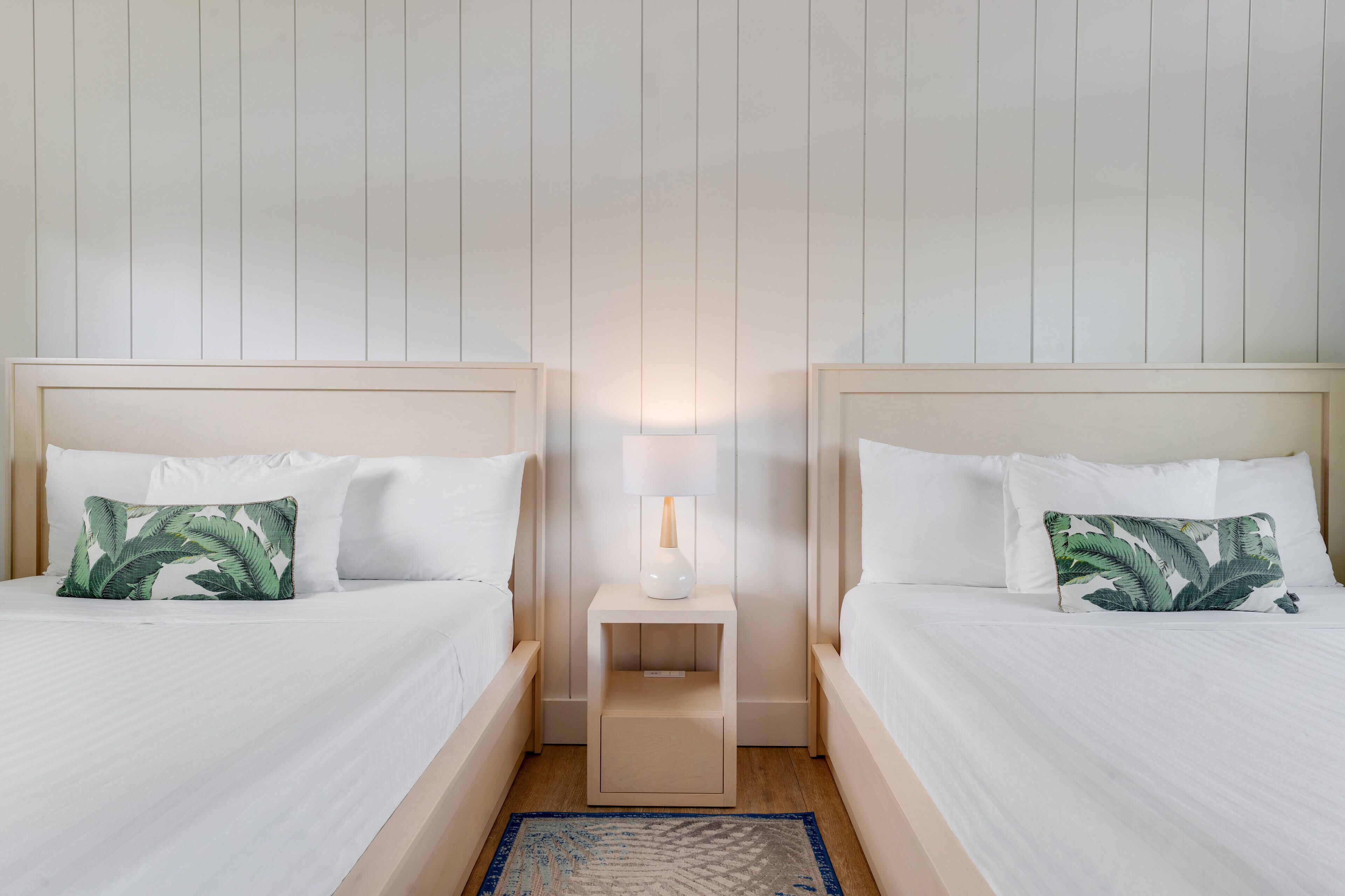 Una habitación de hotel con dos camas, cada una con ropa blanca y una almohada con estampado de hojas verdes. Hay una mesita de noche con una lámpara entre las camas.