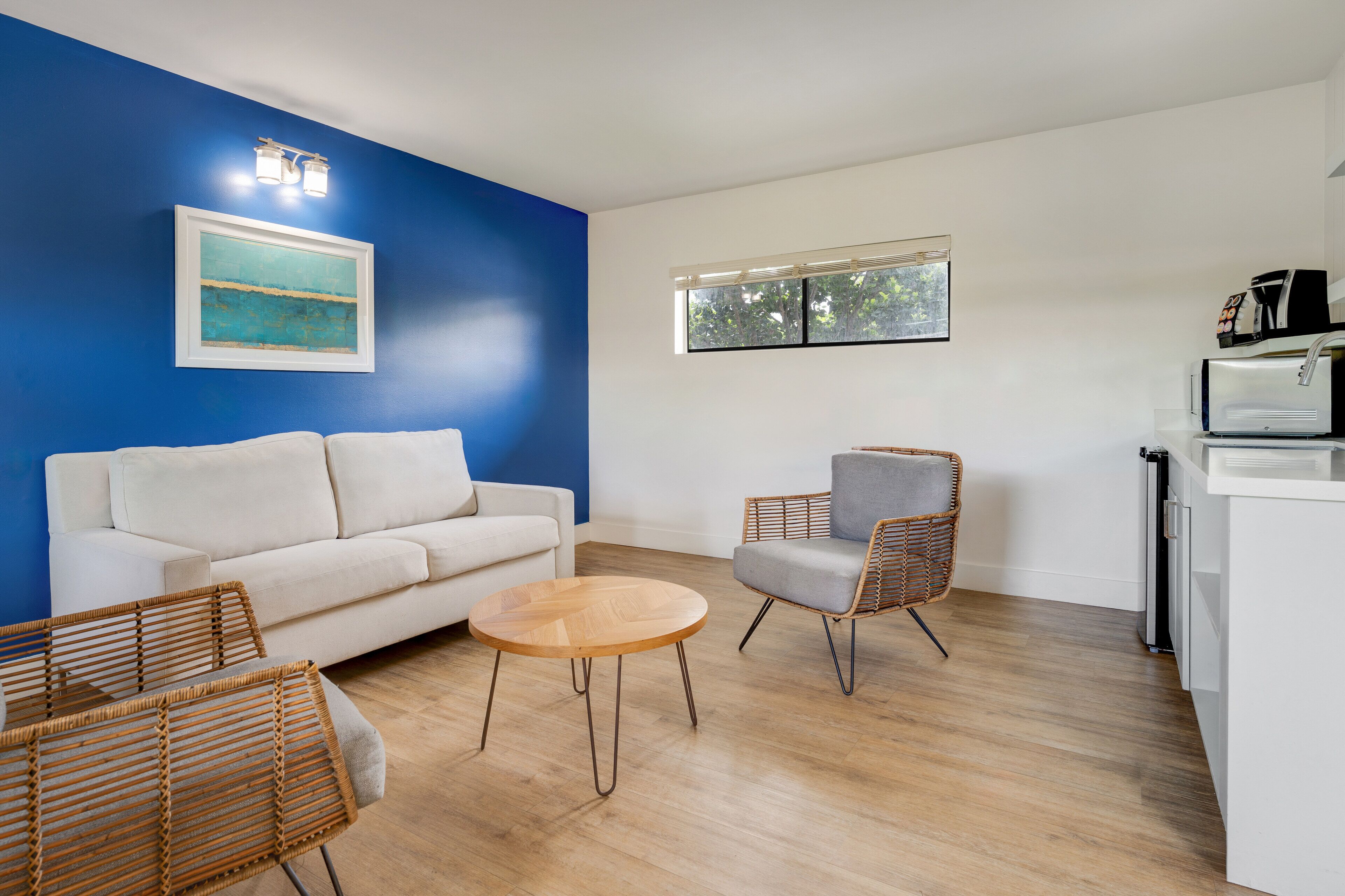 Un salon moderne avec un canapé blanc, une table basse ronde en bois, des fauteuils en osier et un coin cuisine équipé d’un réfrigérateur et d’une machine à café.
