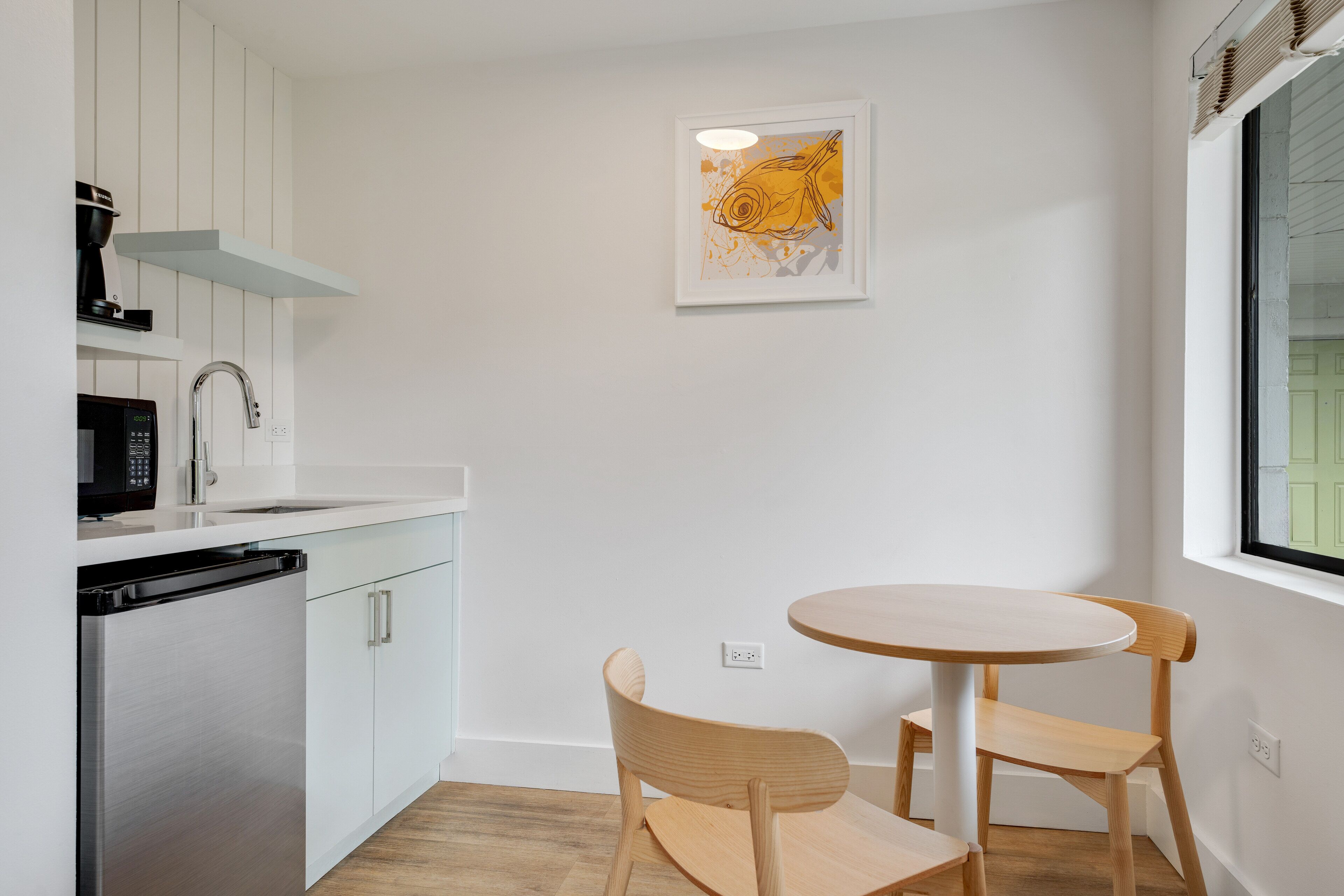 Une cuisine compacte avec des meubles blancs, un réfrigérateur en acier inoxydable, un four à micro-ondes et une machine à café. On y trouve une petite table en bois avec deux chaises et une œuvre d’art encadrée accrochée au mur.