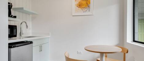 Une cuisine compacte avec des meubles blancs, un réfrigérateur en acier inoxydable, un four à micro-ondes et une machine à café. On y trouve une petite table en bois avec deux chaises et une œuvre d’art encadrée accrochée au mur.