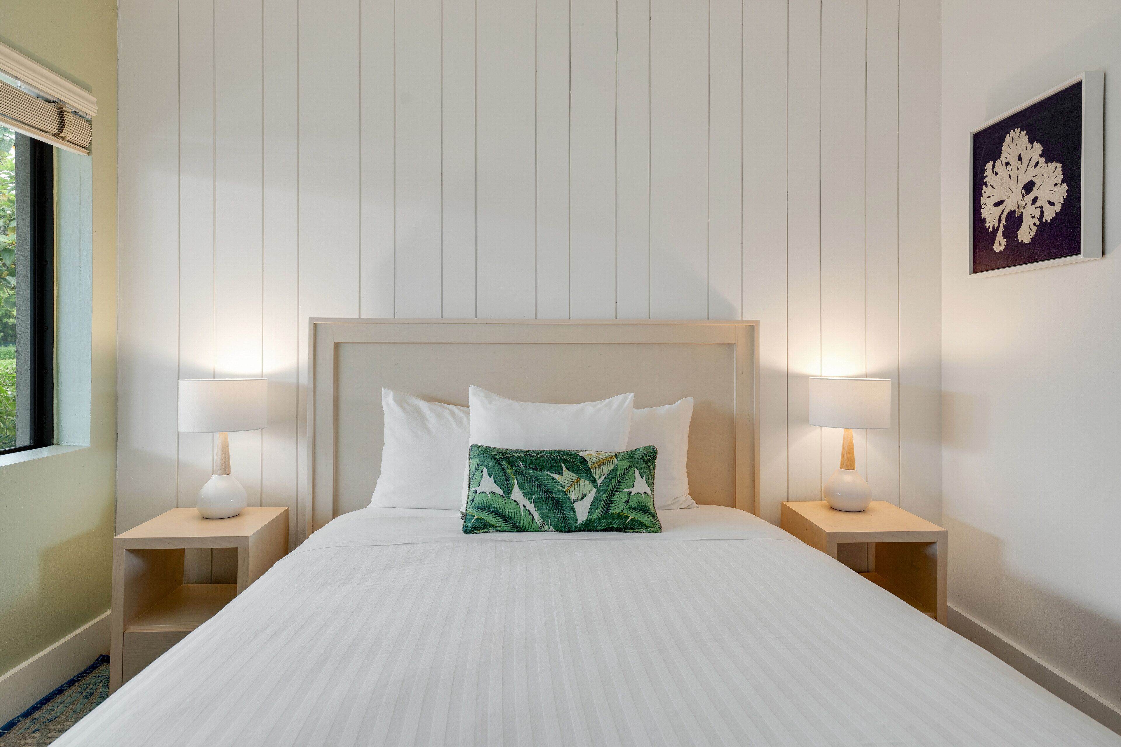 Una cama bien hecha con sábanas blancas, dos mesitas de noche con lámparas y una obra de arte colgada en la pared.