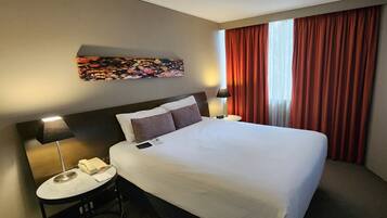 A hotel room with a large bed, a desk with a lamp, a telephone, and a painting on the wall.