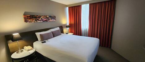 Ein Hotelzimmer mit einem großen Bett, einem Schreibtisch mit Lampe, einem Telefon und einem Bild an der Wand.