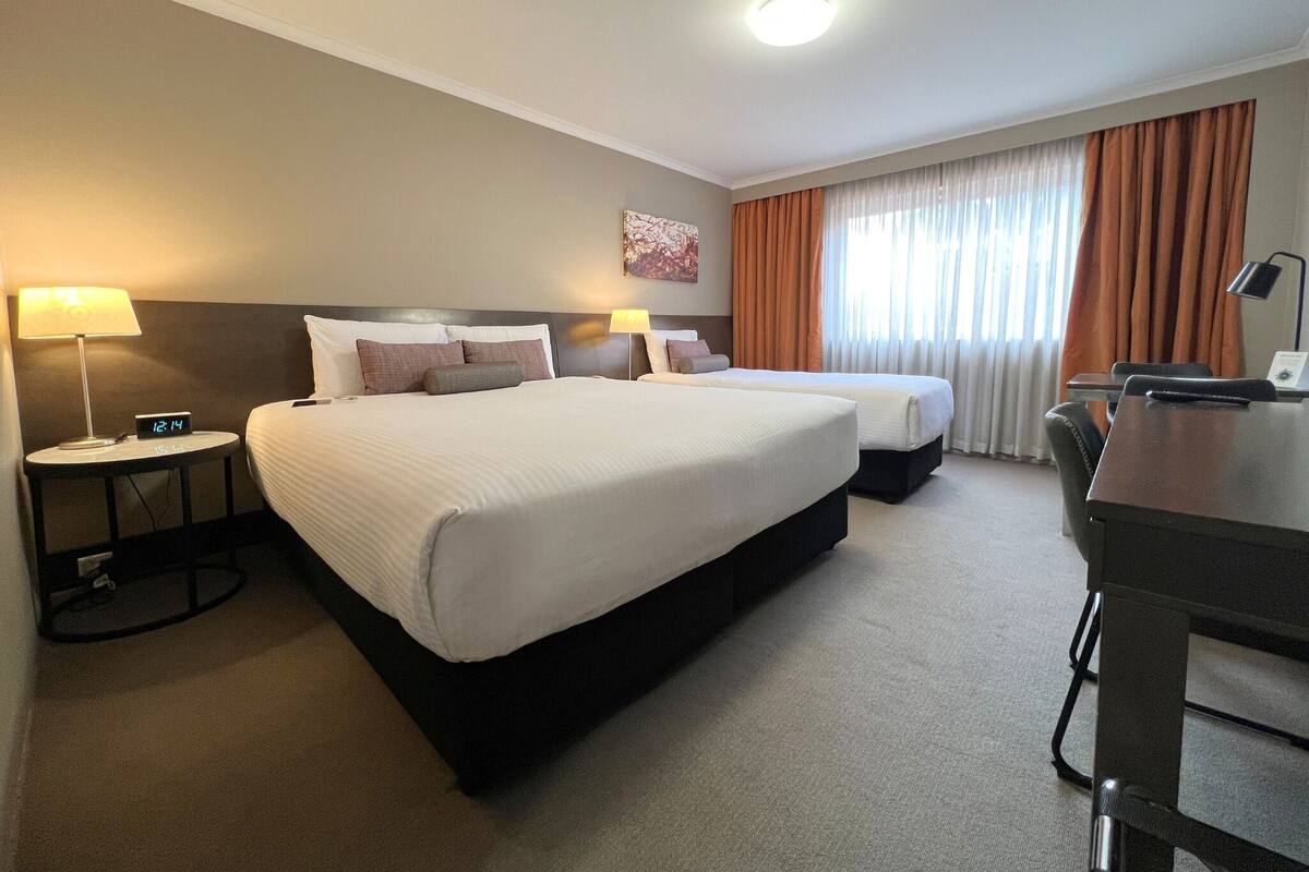A hotel room with two beds, a desk, and a chair. There is a lamp on the left side and a painting on the wall.