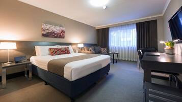 Quarto de hotel com cama grande, escrivaninha, cadeira, televisão e um quadro na parede.