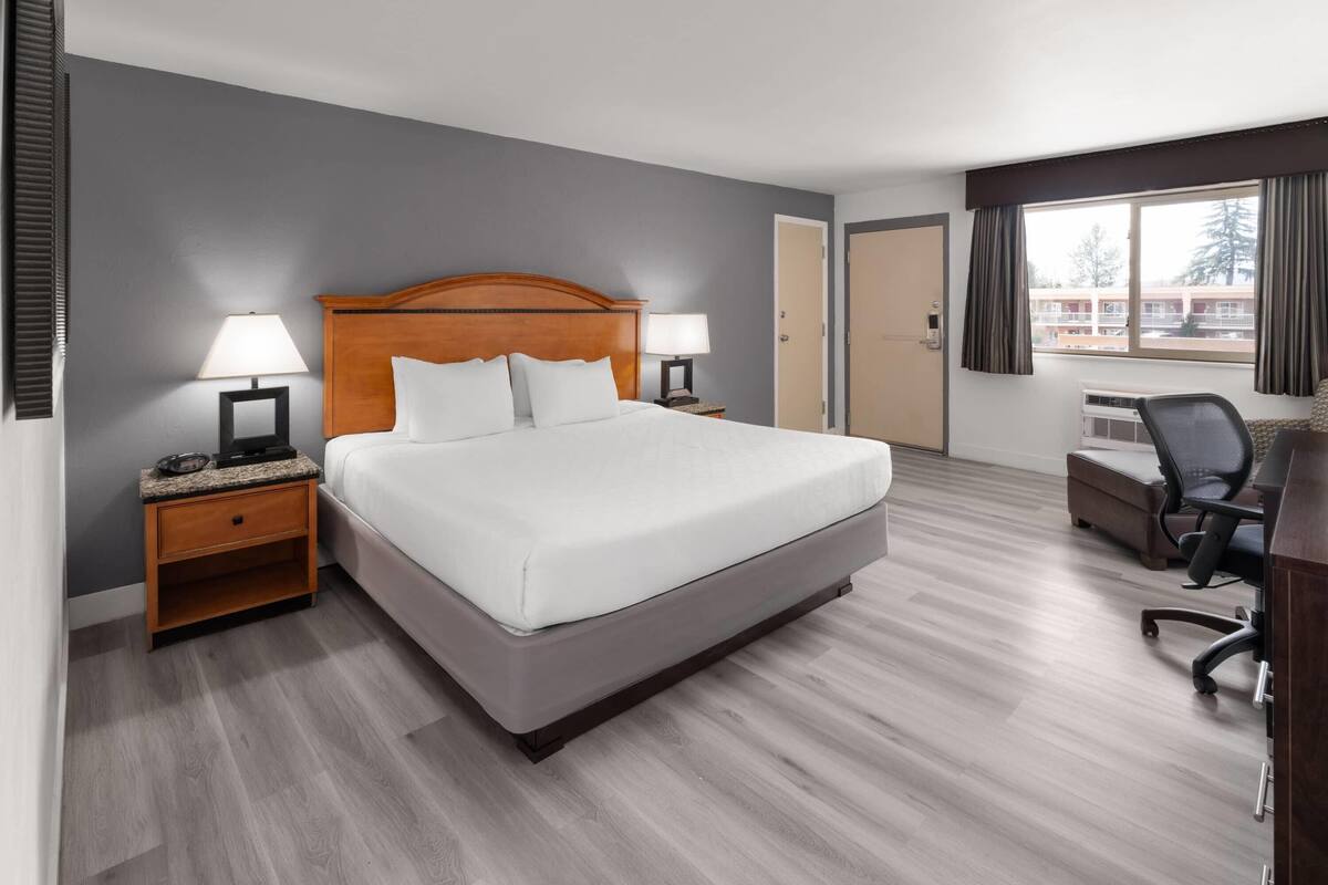 A hotel room with a bed, two bedside tables with lamps, a desk with a chair, and a window with curtains.