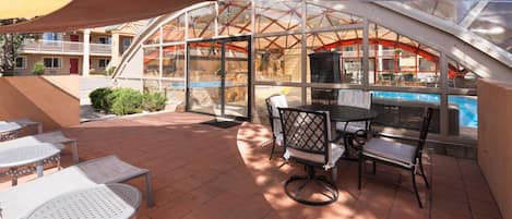 An outdoor patio with tables and chairs, a glass enclosure, and a pool in the background.