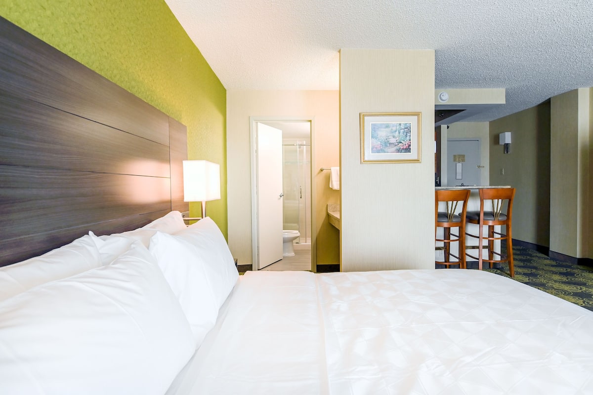 A hotel room with a large bed, a wooden headboard, a desk with two chairs, and a bathroom visible through an open door.