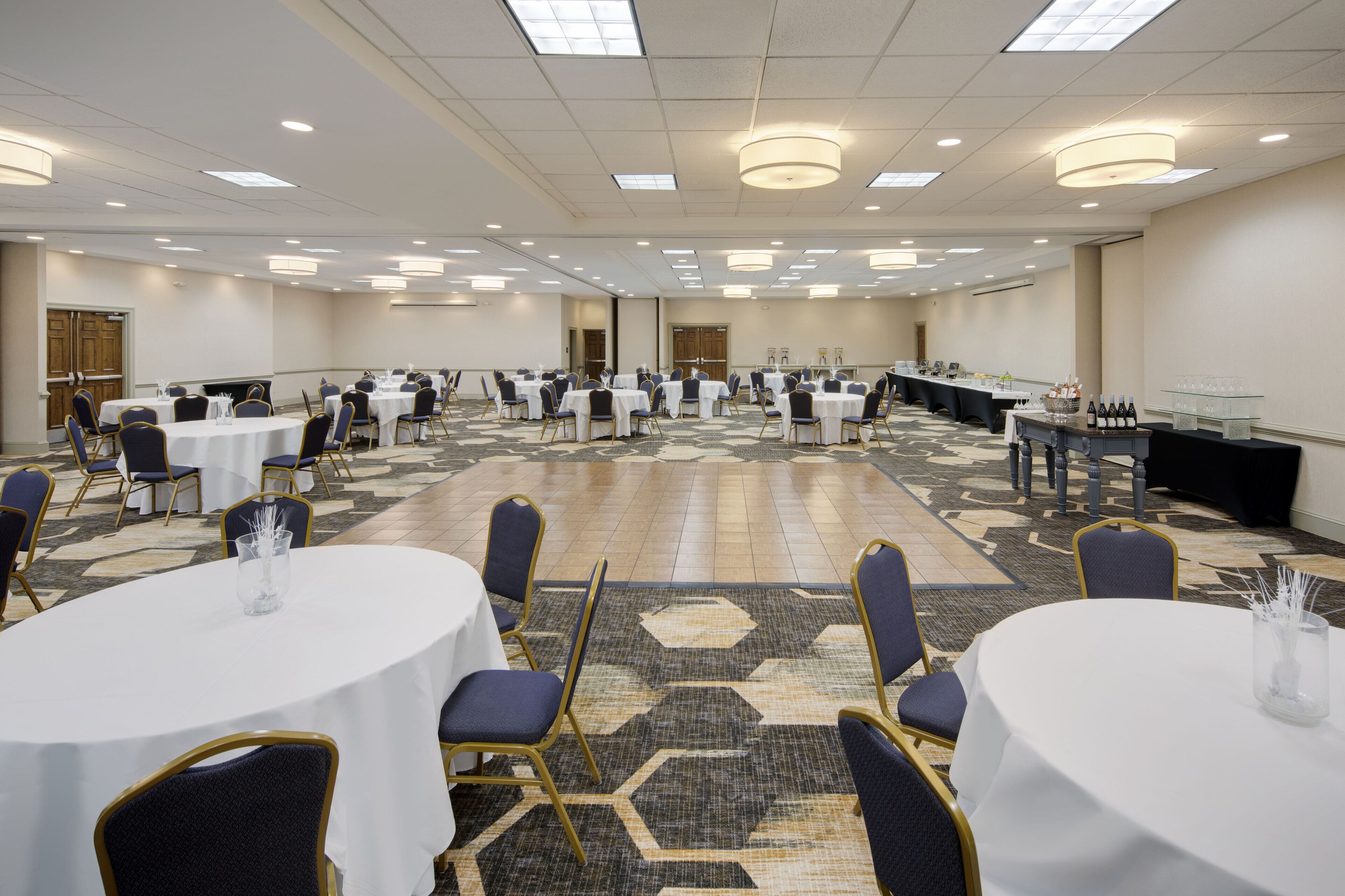 Une grande salle de conférence avec des tables rondes et des chaises, une longue table où est dressé un buffet, et un sol recouvert de moquette.
