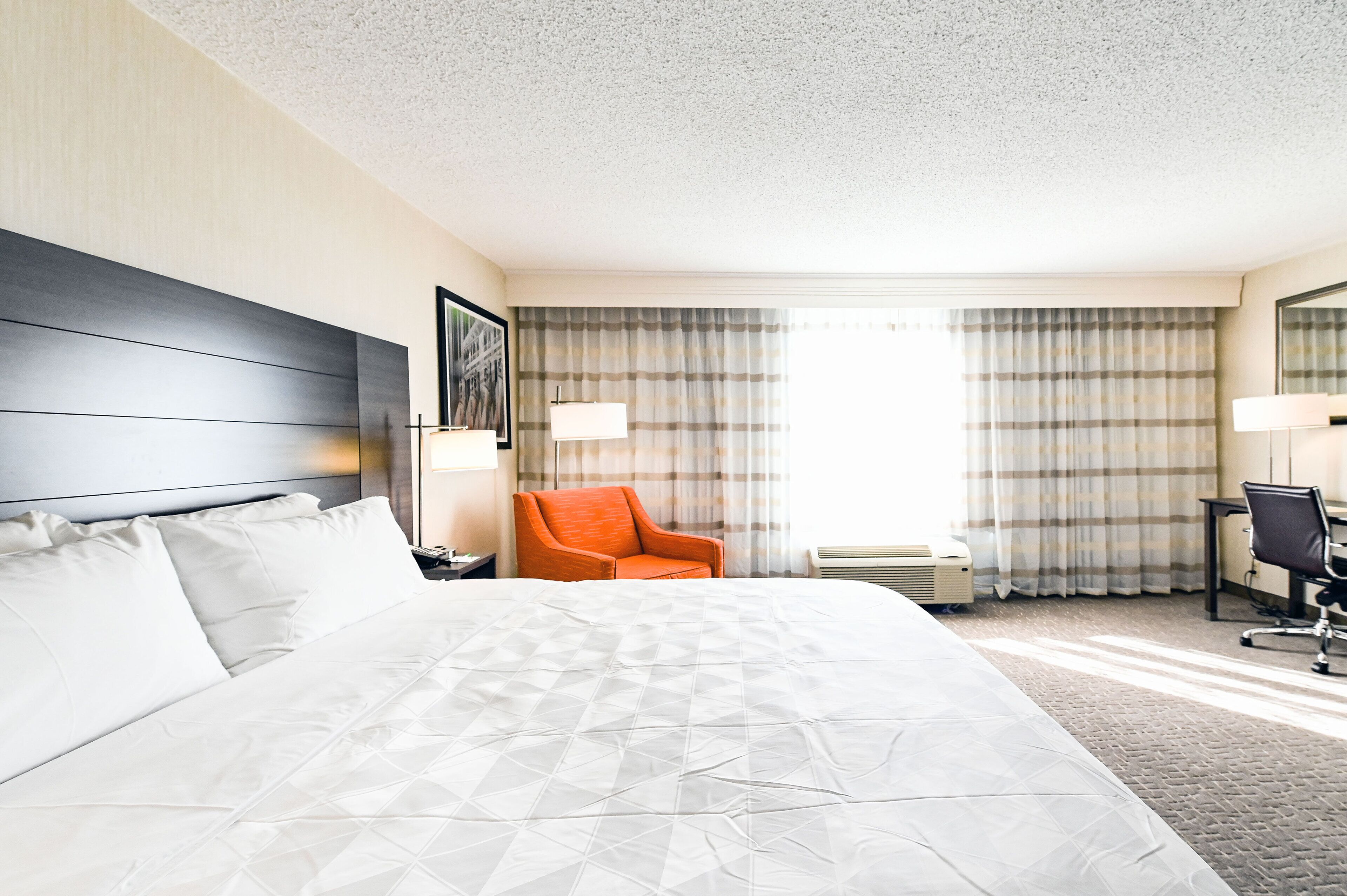 Een hotelkamer met een groot bed, een oranje fauteuil, een bureau en een raam met gordijnen.
