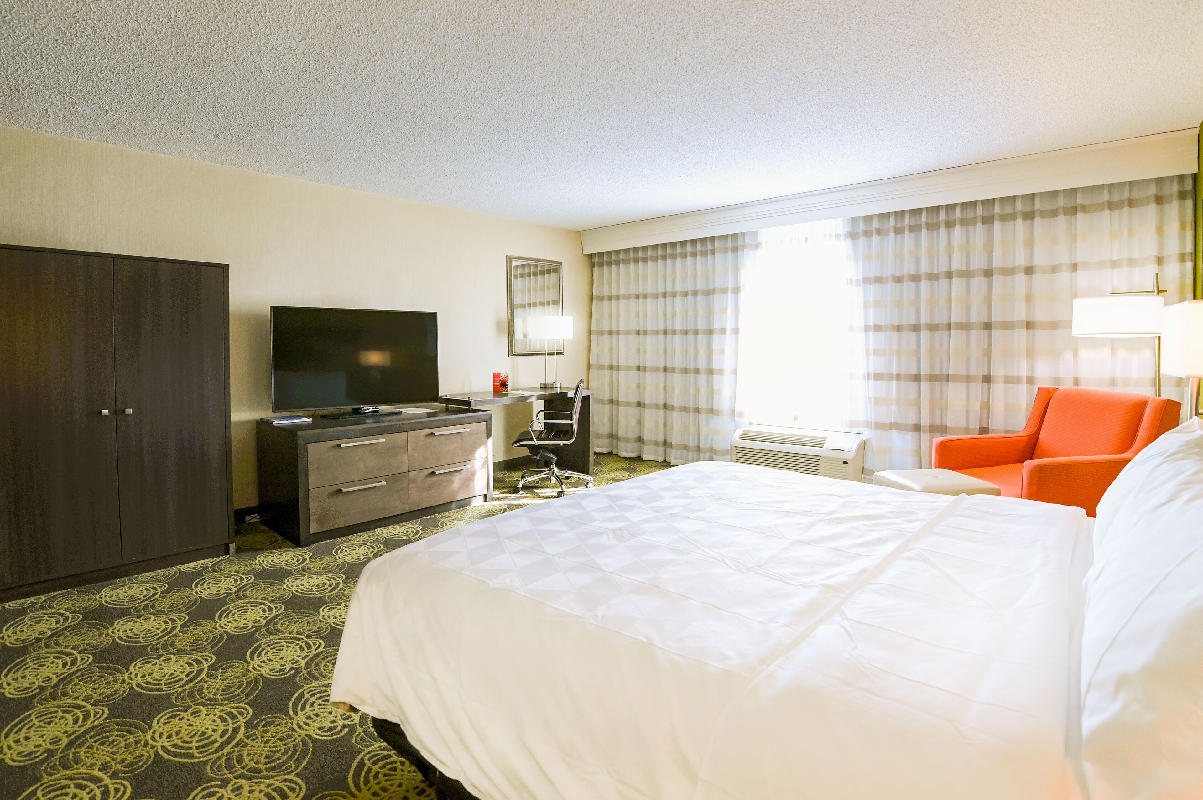 A hotel room with a large bed, a desk with a flat-screen TV, a chair, and a wardrobe.