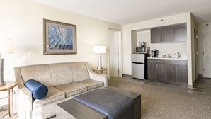 A hotel room with a sofa, ottoman, and a small table with a lamp. There is a kitchenette with a microwave, coffee maker, and cabinets. A framed picture hangs on the wall. - Holiday Inn Charlottesville-Univ Area by IHG (Charlottesville)