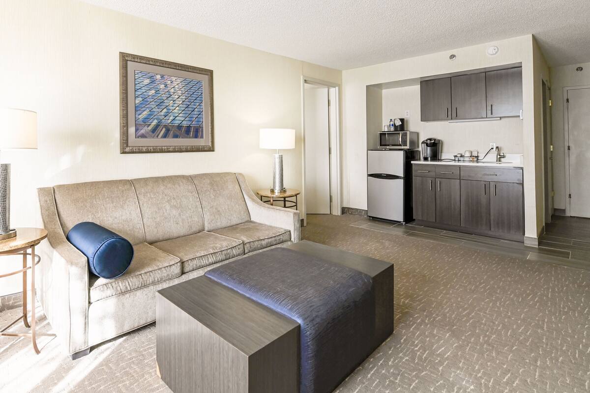 A hotel room with a sofa, ottoman, and a small table with a lamp. There is a kitchenette with a microwave, coffee maker, and cabinets. A framed picture hangs on the wall.