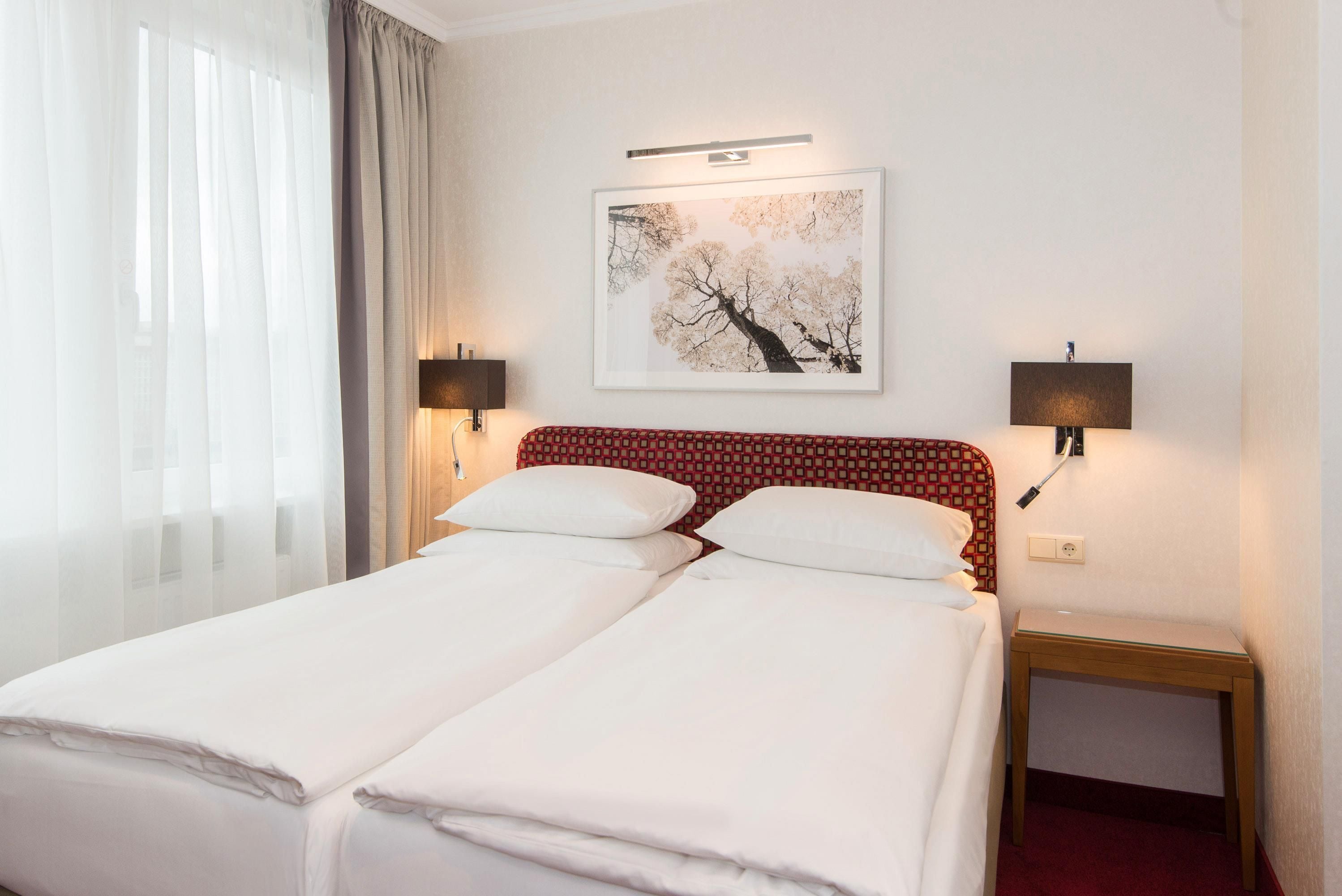 A hotel room with two beds, a nightstand, a lamp, a wall-mounted picture, and a window with curtains.