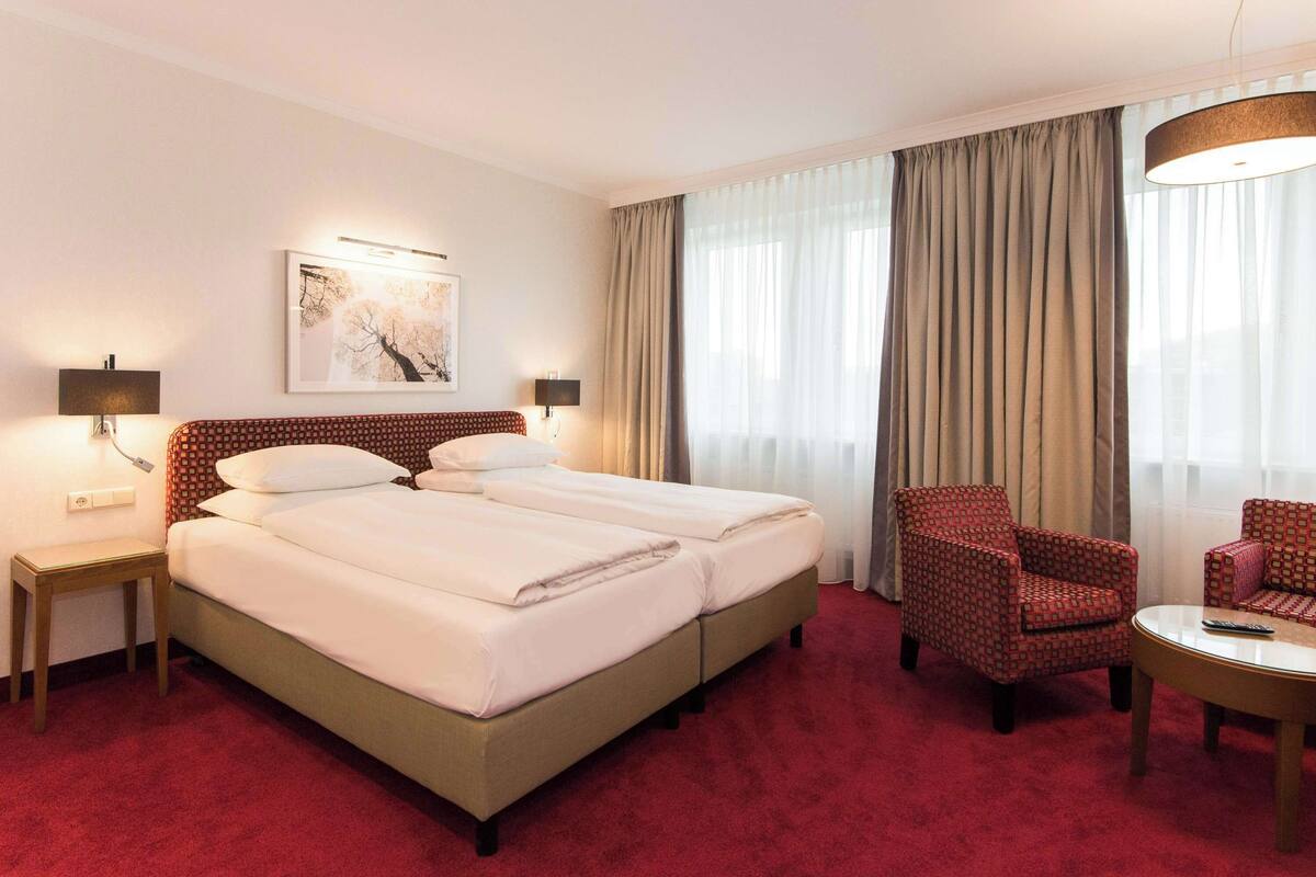 A hotel room with two beds, a red armchair, a small table, and a painting on the wall.