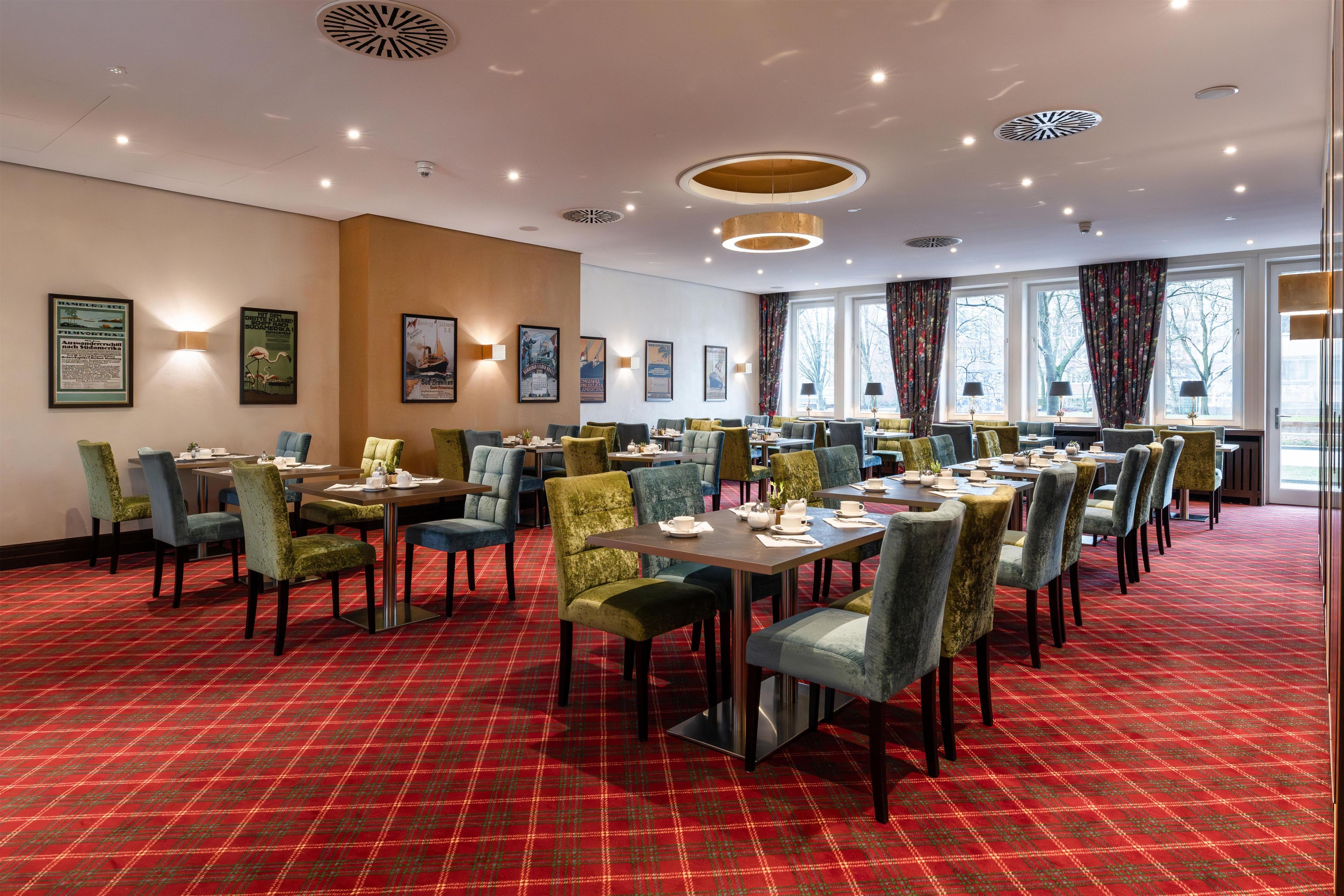 A hotel dining area with tables and chairs, framed pictures on the walls, and large windows with curtains.