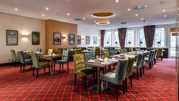 A hotel dining area with tables and chairs, framed pictures on the walls, and large windows with curtains.