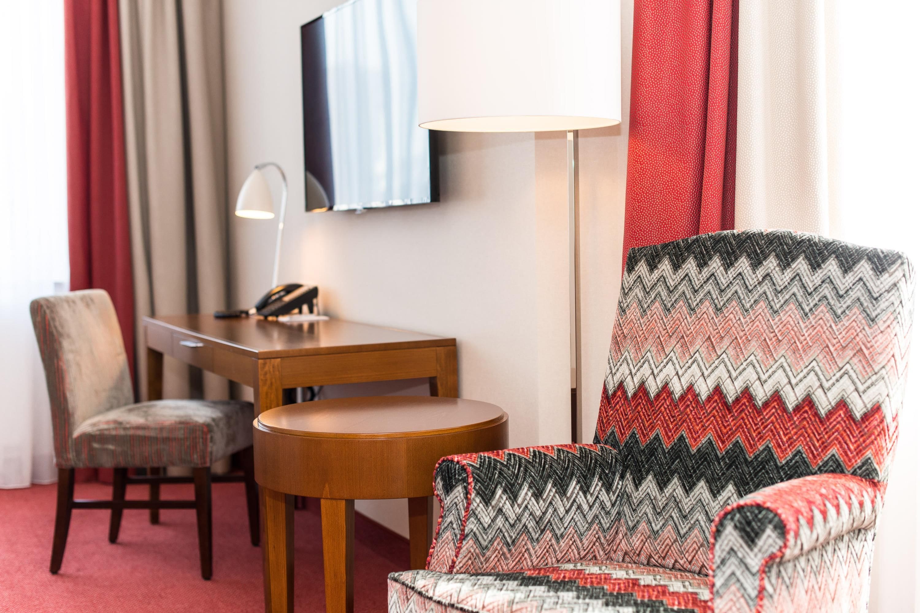 Ein Hotelzimmer mit Holzschreibtisch, einem Stuhl, einem runden Tisch, einem gemusterten Sessel und einer Lampe.