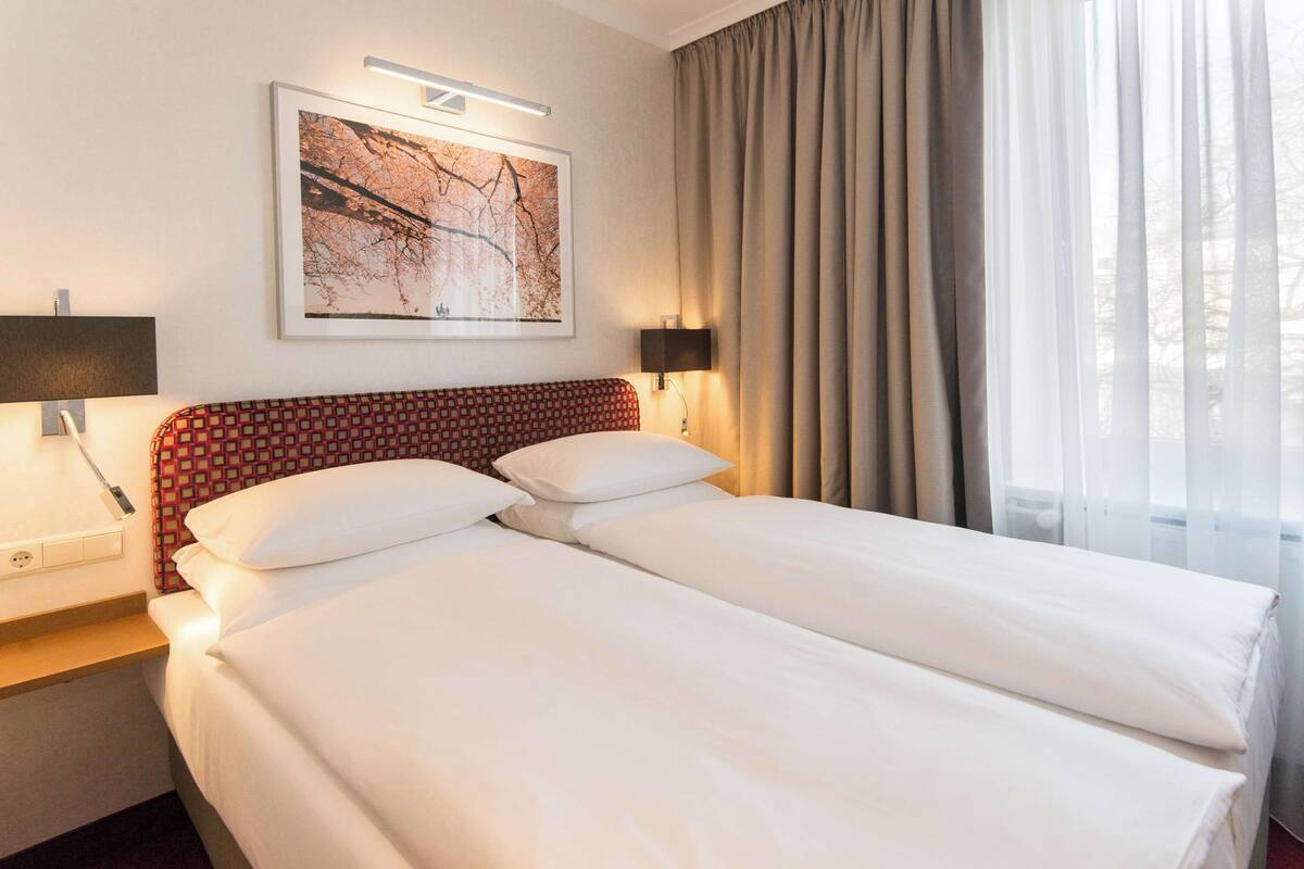 A hotel room with a double bed, a nightstand, a lamp, a framed picture on the wall, and a window with curtains.