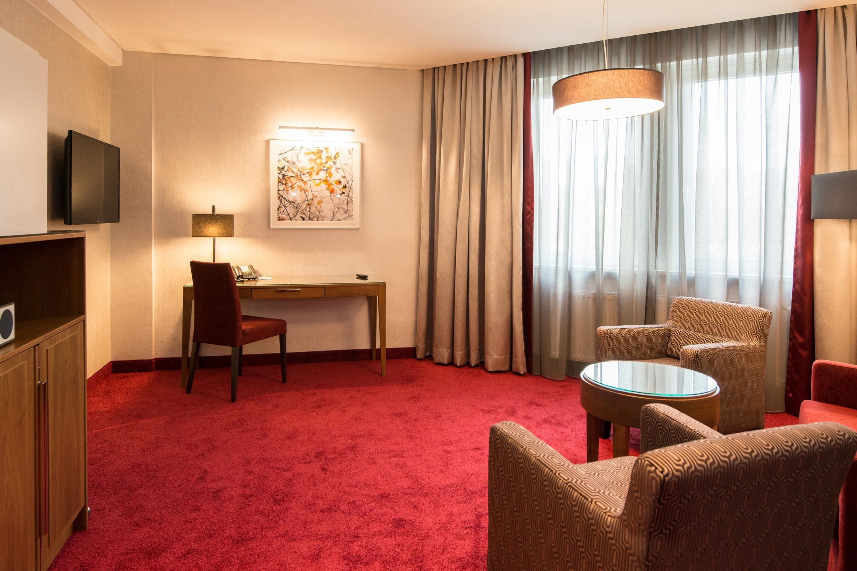 A hotel room with a red carpet, a desk with a chair, a television, a painting, a window with curtains, and a patterned armchair.