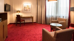 Habitación de hotel con alfombra roja, un escritorio con silla, televisor, un cuadro, una ventana con cortinas y un sillón estampado.