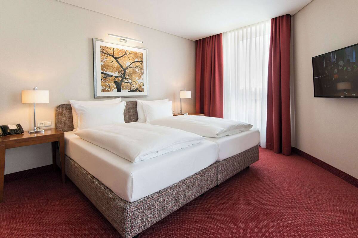 A hotel room with a double bed, two bedside tables with lamps, a large framed picture on the wall, and a TV mounted on the wall.